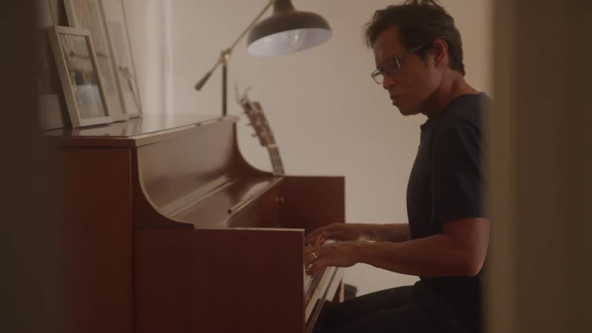 A man wearing glasses plays an upright piano in a warmly lit room