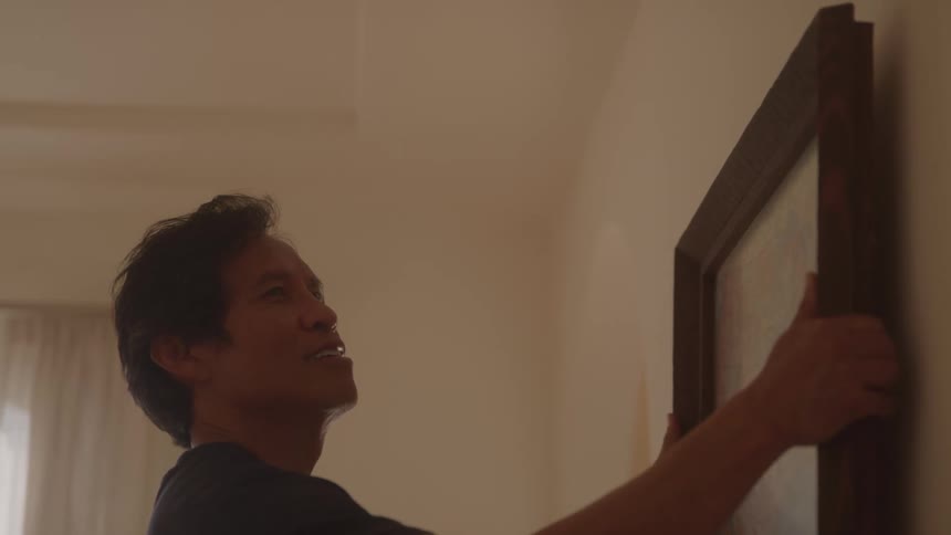 A man carefully picks up a framed picture from a stack against the wall and hangs it