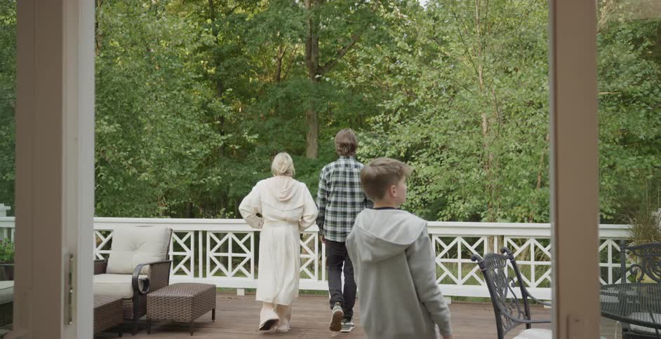 A woman a man and a child stand on a wooden deck with white railings looking out at a dense forest under a bright sky