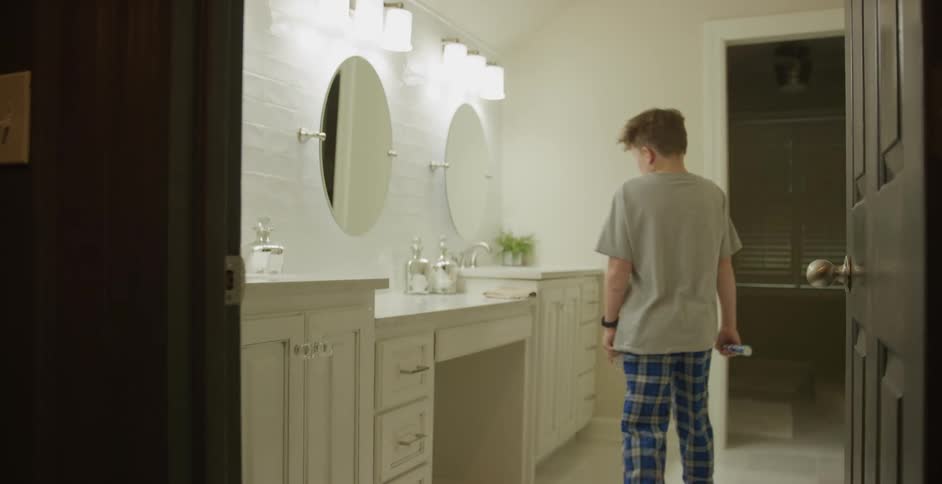 A child enters the bathroom and stands at the sink reflected in the mirror as he reaches for the faucet to brush his teeth
