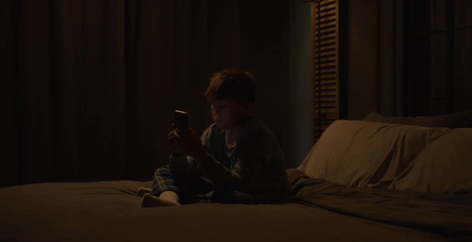 A young boy sits on a bed before sleep looking bored while using a smartphone