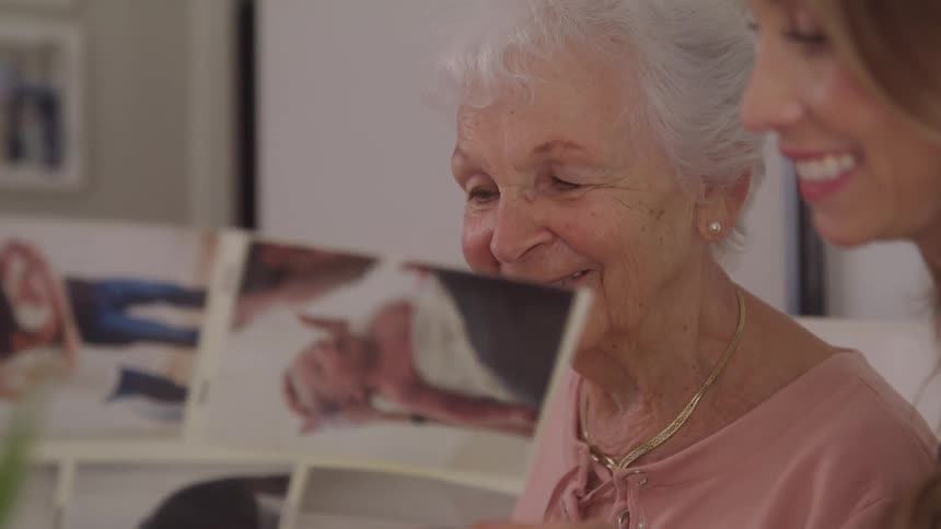 An elderly woman in a pink top and gold necklace sits beside a young woman as they look through a photo album