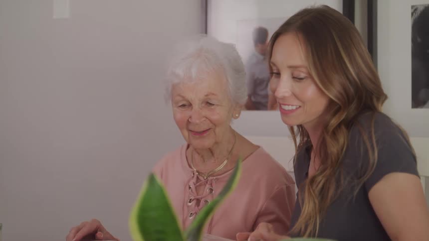 Two women one young and the other elderly sit indoors smiling as they go through a magazine together