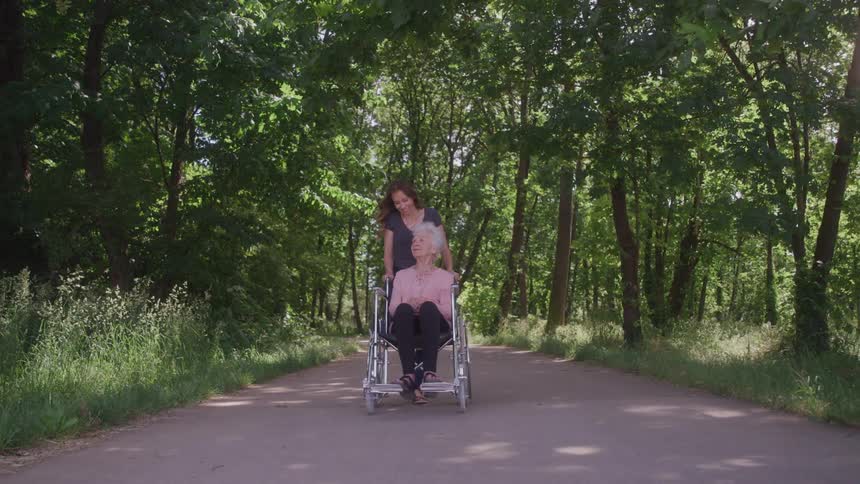A woman pushes an elderly woman in a wheelchair along a public park walkway around midday