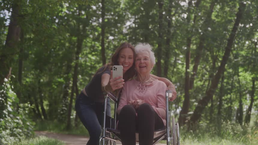 Young woman and elderly woman in wheelchair take a selfie on a forest path