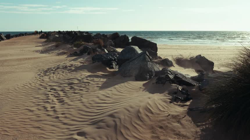 Wind blown sand patterns and large rocks line a quiet beachside pathway under clear skies