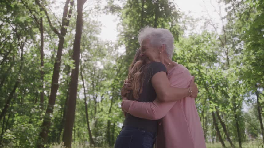 An older woman with white hair embraces a younger woman with long wavy hair both smiling in a sunlit outdoor setting surrounded by trees