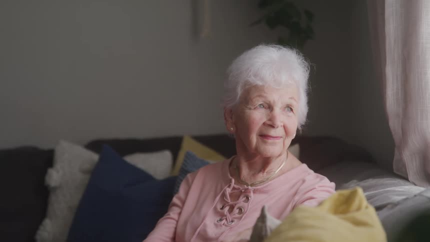 Close up portrait of an elderly woman looking out the window while sitting on a couch in a living room during daytime