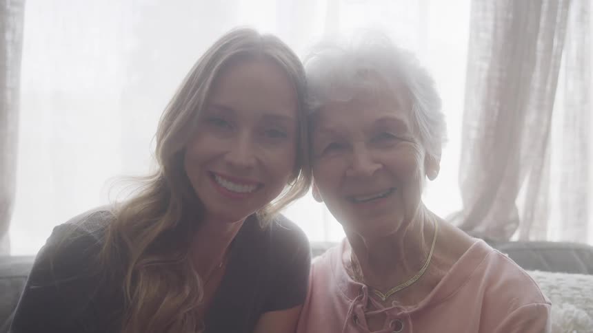 Close up portrait of a young woman and elderly woman sitting and smiling together on a couch in the living room during daytime