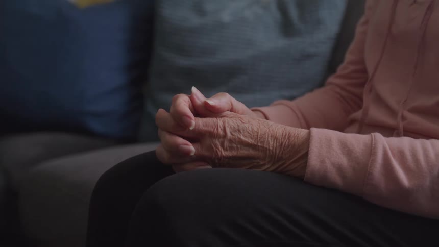 A close up view shows the hands of an older woman clasped together resting on her lap against a dark blurred background