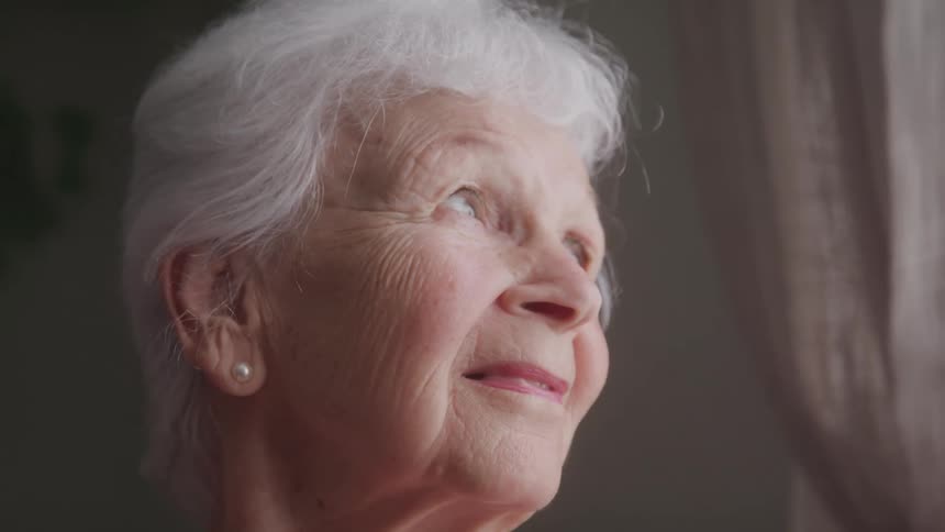 Close up portrait of an elderly woman smiling while looking out of the window in a room during daytime
