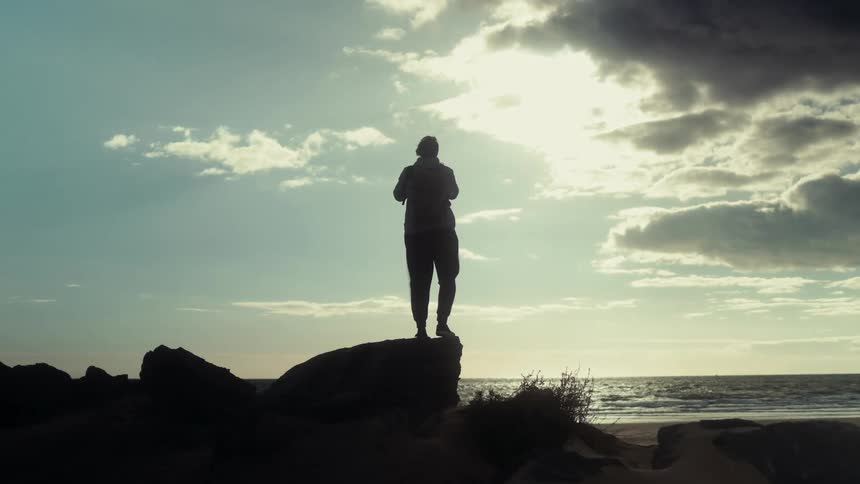 A person stands on a large rock at the beach silhouetted against a dramatic sky during sunset