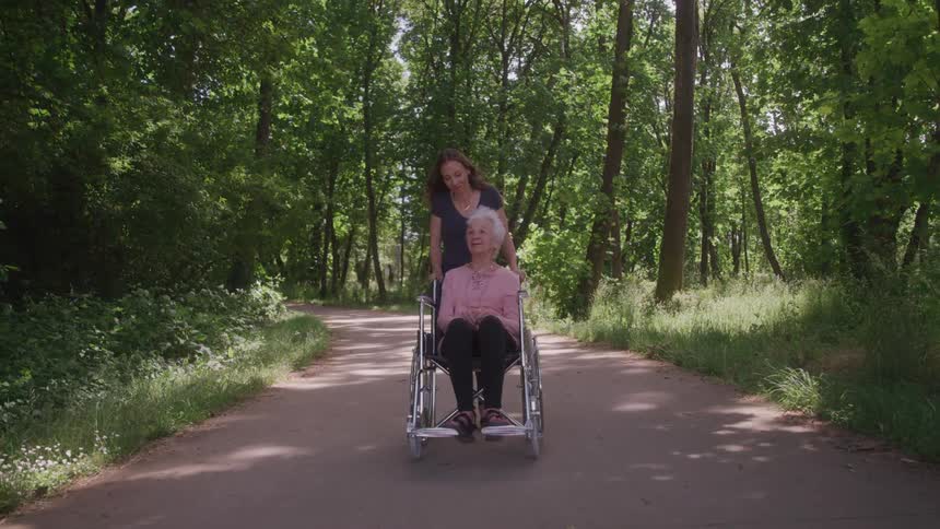 Woman pushes elderly lady in wheelchair along forest path