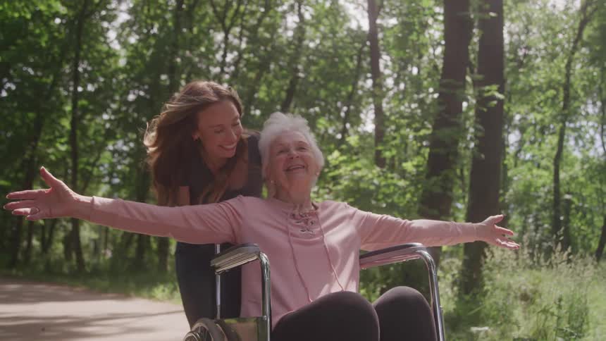 A young woman pushing an elderly woman in a wheelchair who has her arms open and is smiling happily