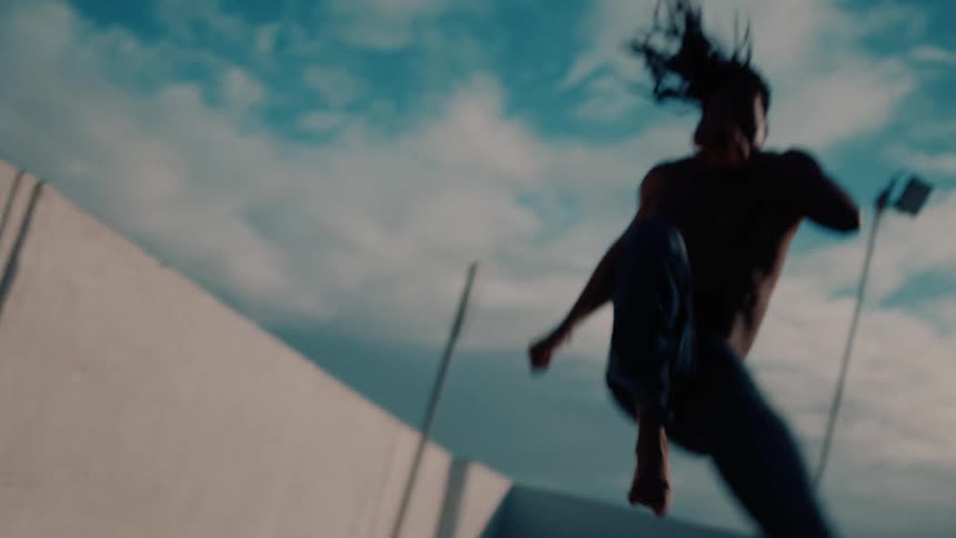 A shirtless man shows off martial arts moves next to a white building in a city