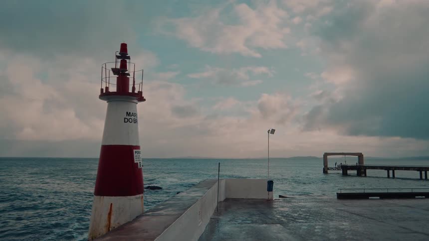 A lighthouse stands at the edge of a pier overlooking the sea under a partly cloudy sky