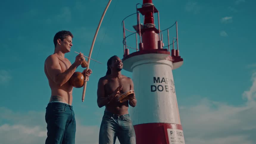 Two shirtless men play traditional instruments next to a lighthouse under a clear blue sky