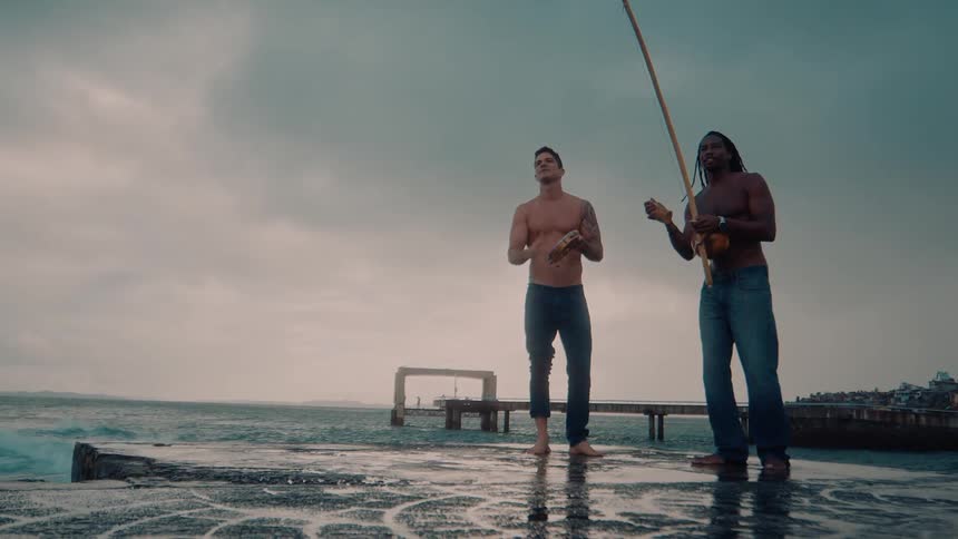 Two men stand on a wet pier by the ocean one playing a tambourine while the other plays percussion beside him
