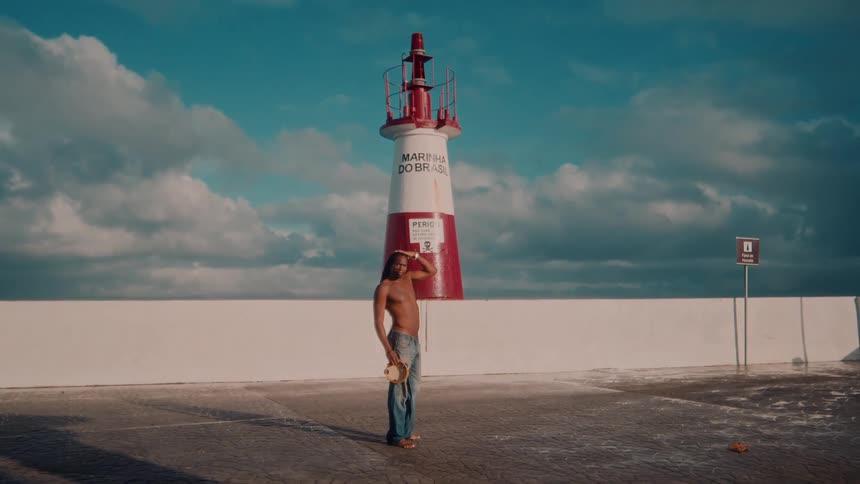 A man holding a tambourine stands on a sunny seaside promenade in front of a red and white lighthouse under a cloudy blue sky