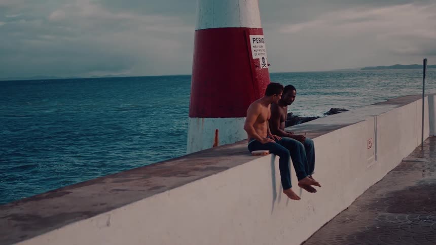 Two men in jeans and no shirts sitting next to a lighthouse on the seashore