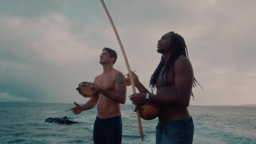 A man playing a tambourine and another man playing a berimbau at the beach shore