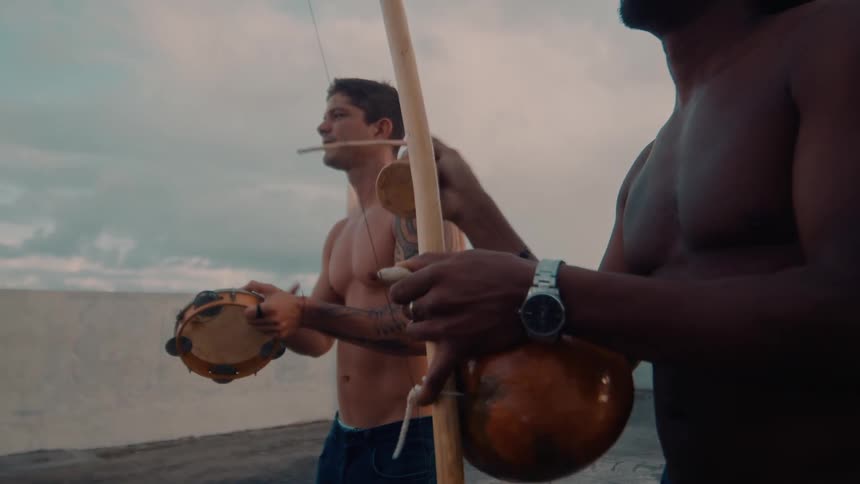 Two shirtless men playing berimbau at a seaside fort