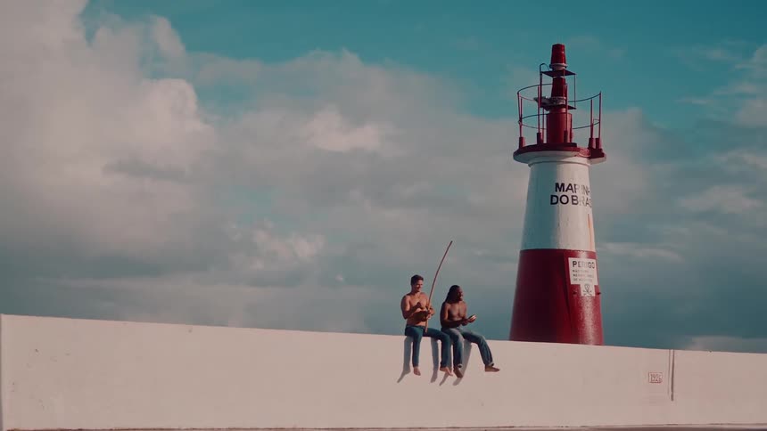 Two shirtless men are seated on a white wall near a red and white lighthouse with water splashing under a partly cloudy sky