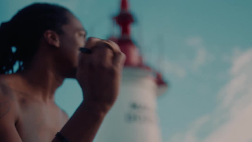 A young man with dreadlocks shakes a musical instrument beside a lighthouse