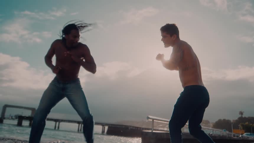 Two men in jeans in a seaside setting fighting capoeira