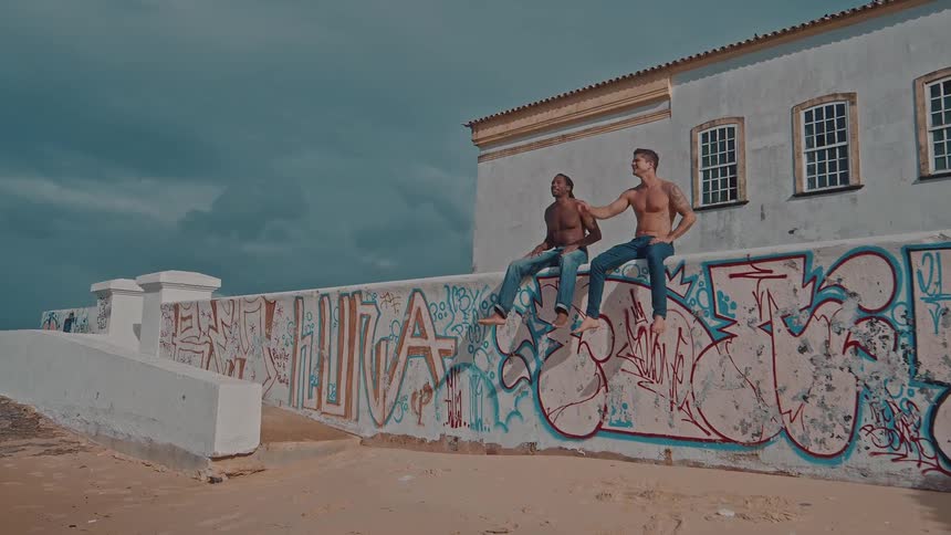 Two shirtless men chat sitting on a graffiticovered wall