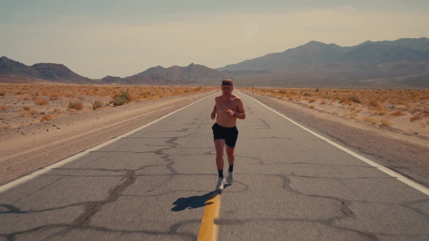 A shirtless man runs down a cracked desert highway with mountains in the distance