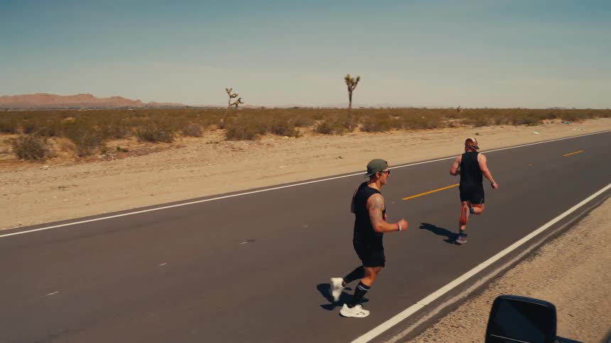 A young man runs along the side of a highway under the sun he shakes hands with another man who then starts running as the first man stops a car drives by them