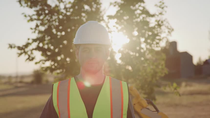 A man wearing a white hard hat and a safety vest stands outdoors with sunlight filtering through trees behind him