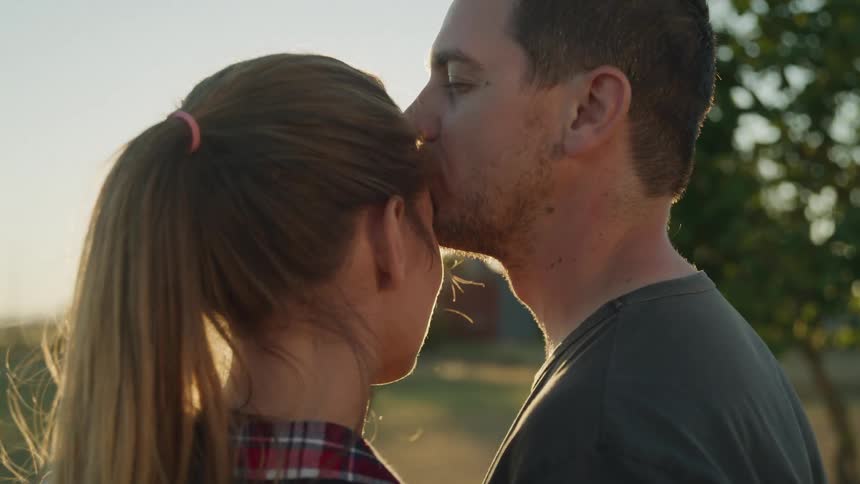 Man and woman stand looking towards the sun before the man kisses the woman on her forehead