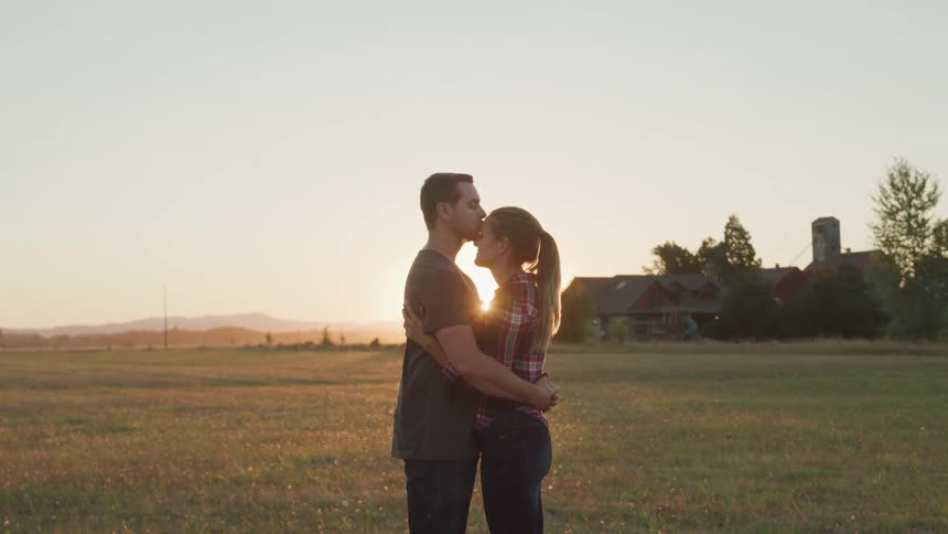 Man hugs and kisses a womans forehead while standing on a field at sunset