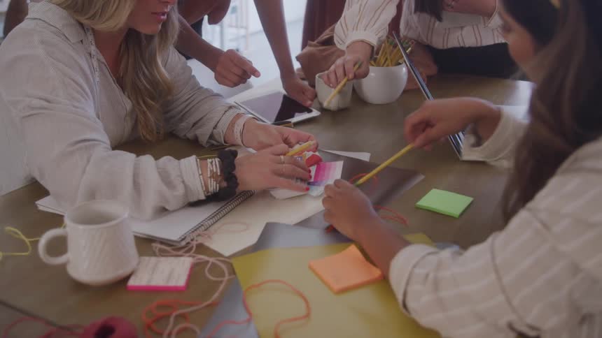 Several women are gathered around a table cluttered with various craft supplies including pencils sticky notes and yarn