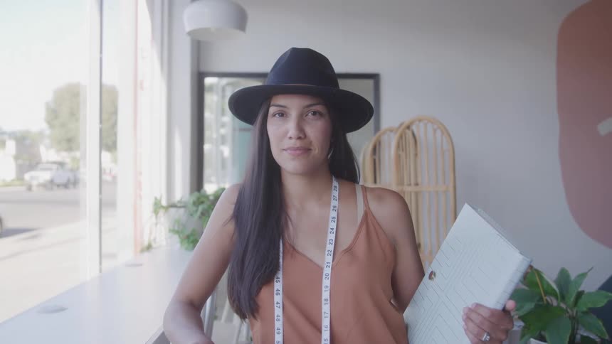 A confident woman stands indoors with a notebook and measuring tape around her neck