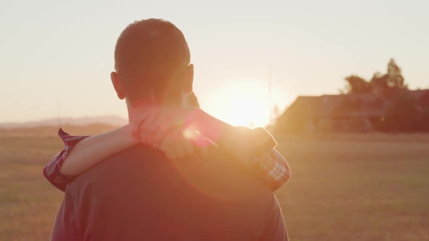A woman with a ponytail wearing a plaid shirt embraces a man and smiles at him in a field at sunset