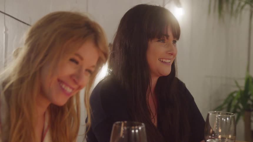 Two women smile and laugh while sitting at a warmly lit dinner table