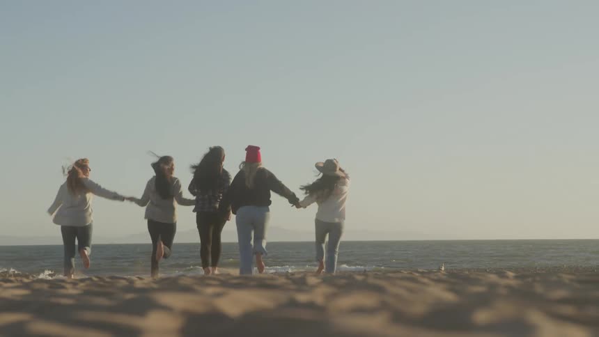Five women run barefoot toward the ocean holding hands and laughing