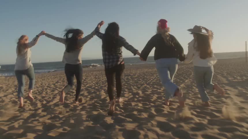 Five women run barefoot along a sunny beach holding hands and kicking up sand