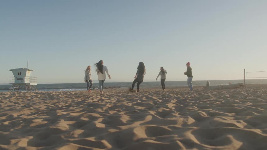 A group of friends play soccer on a sandy beach near a lifeguard stand with the ocean and a clear sky