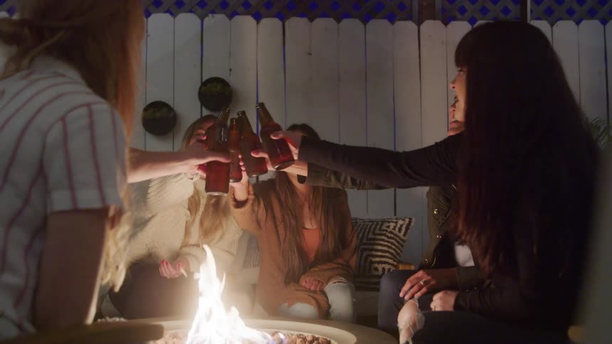 Friends cheer with drinks around a cozy outdoor firepit at night