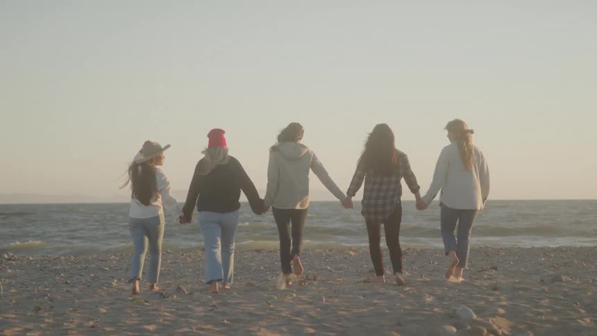 Five women walks towards the beach water while holdings hands together at sunset
