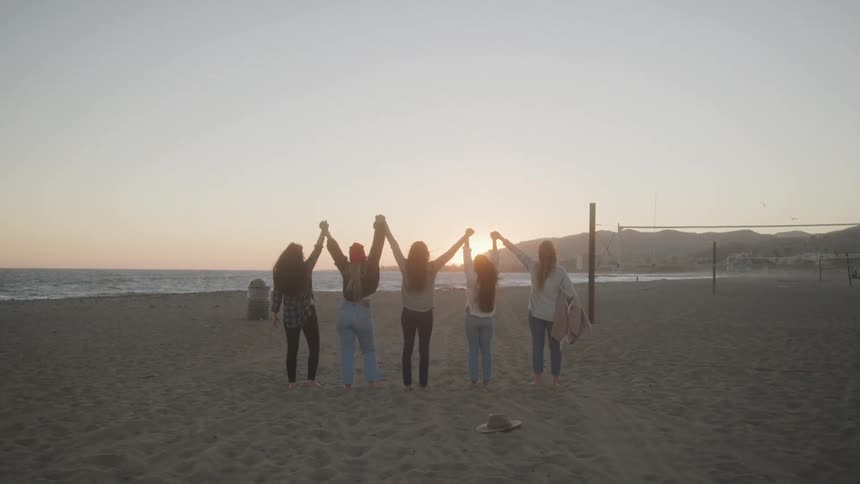 A group of friends stand barefoot on a beach at sunset holding hands with arms raised against the glowing horizon