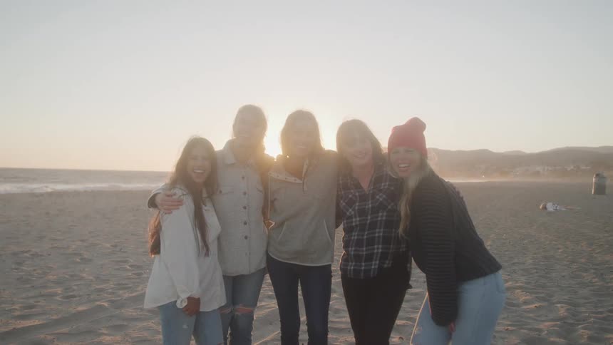 Five women smiling while standing close together at the beach at sunset