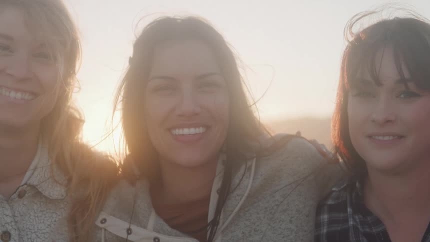 Three smiling women stand close together their faces illuminated by a bright warm sun flare
