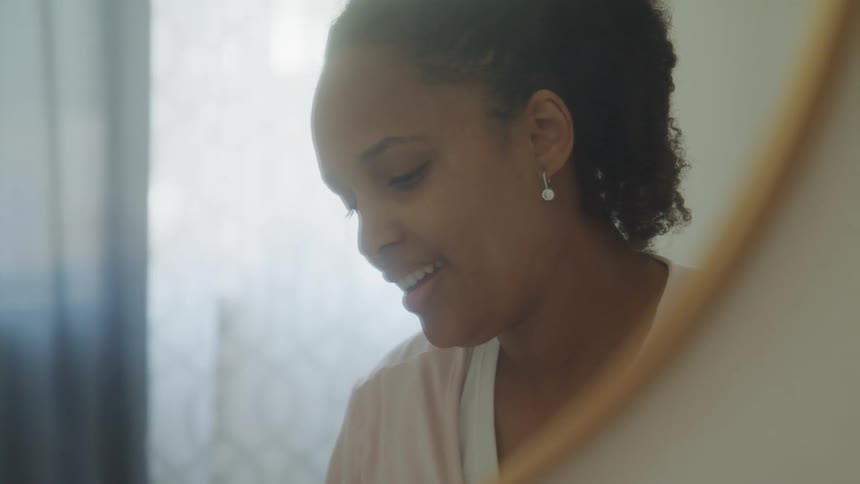 A woman with curly hair wearing a white top and pink robe with a pearl earring stands in front of a mirror in a softly lit room