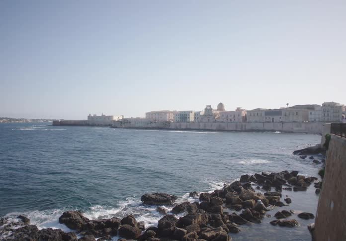 A scenic view shows the historic stone buildings of a coastal city bordering the blue sea with a rocky shoreline in the foreground