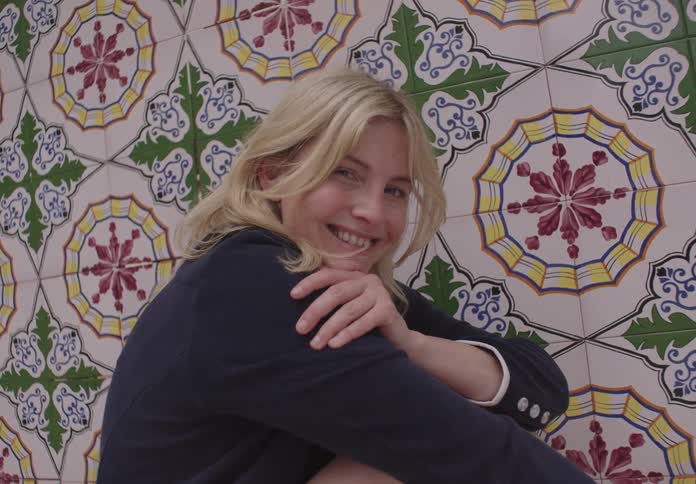 A young woman sits beside a tiled wall smiling continuously
